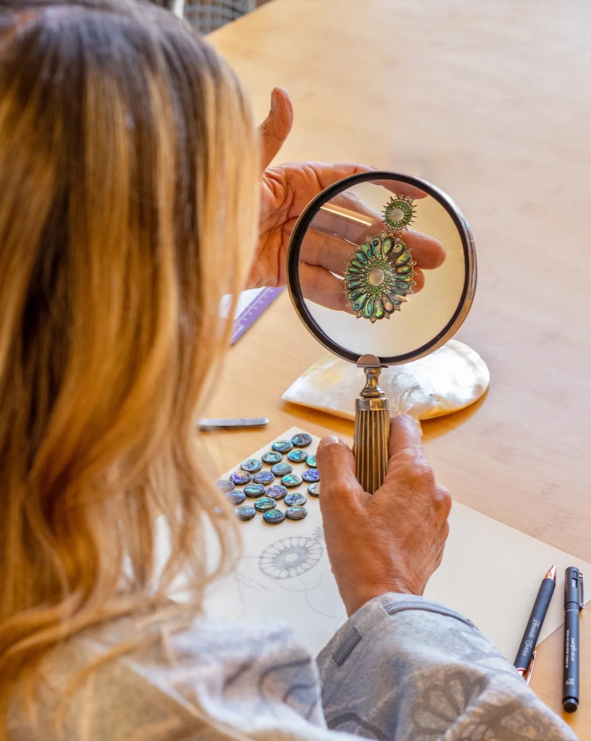 A designer examines a jewel through a magnifying glass at a work table with sketches and coloured stones