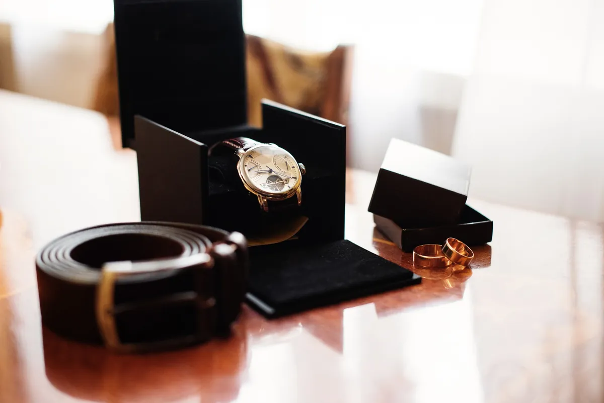 A dress watch in its presentation box next to a leather belt and two rings